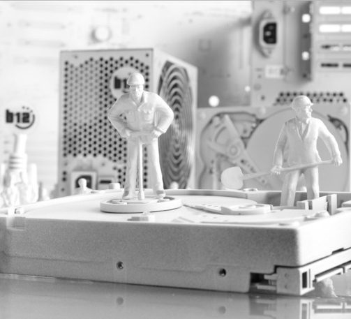 IT-Werkstatt Zwei Figuren stehen auf einer Computerfestplatte vor technischen Bauteilen.